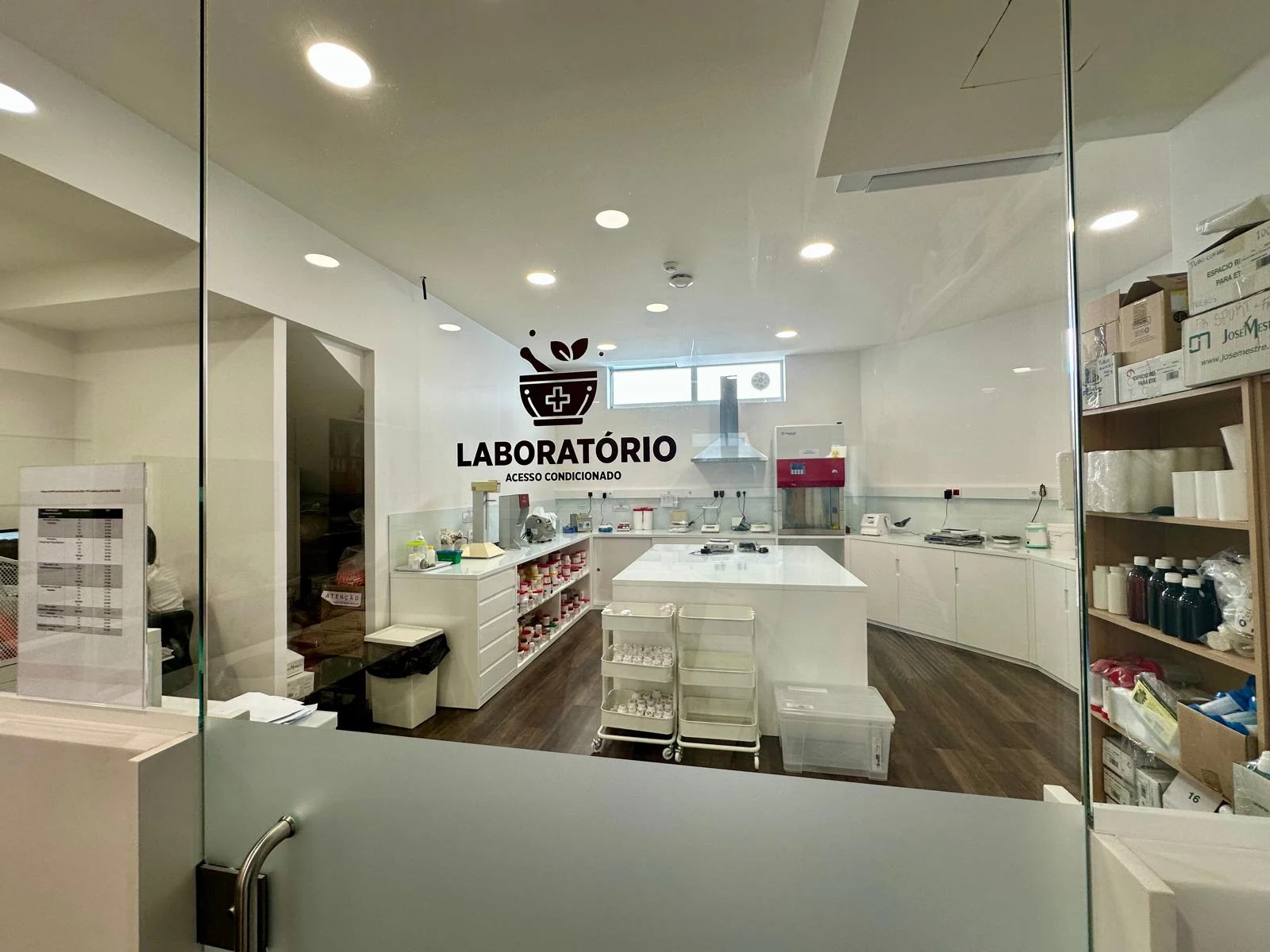Entrada envidraçada do laboratório de manipulados, com bancadas e equipamentos no interior